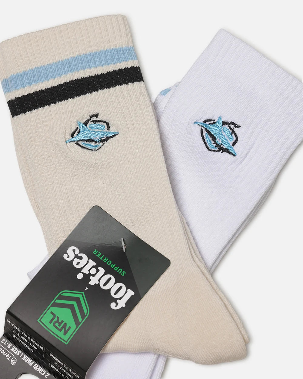 Two pairs of crew socks from Footies x NRL, featuring the Cronulla Sharks logo. One pair is cream/beige with light blue and black stripes, and the other is white.