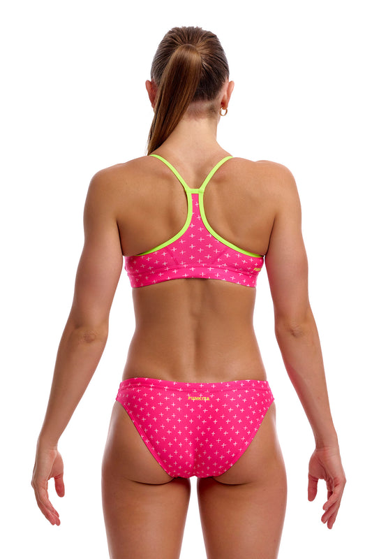 Rear view of a female model standing against a solid white background wearing the Funkita "Pink Proton" Women's Swim Crop Top and matching bottoms, highlighting the lime green racerback single-bind shoulder straps and bright pink geometric print.