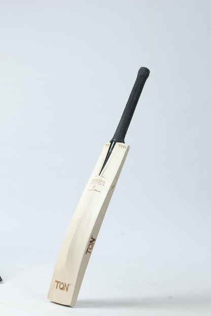A wooden cricket bat with a black textured handle, standing upright against a light gray studio background. The brand name 'TON' is printed on the blade.
