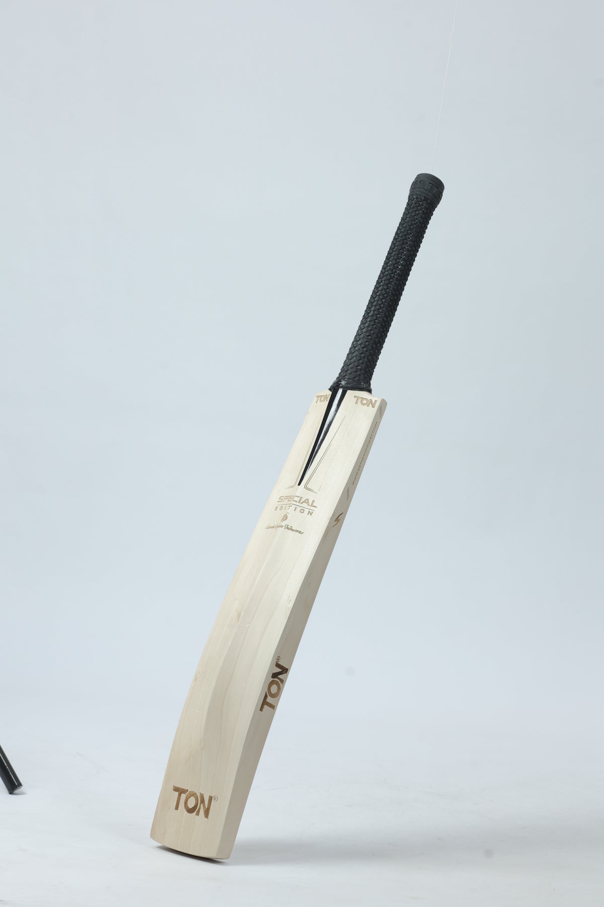 A wooden cricket bat with a black textured grip handle, standing upright against a light gray studio background. The bat features the brand name 'TON' printed on the blade.