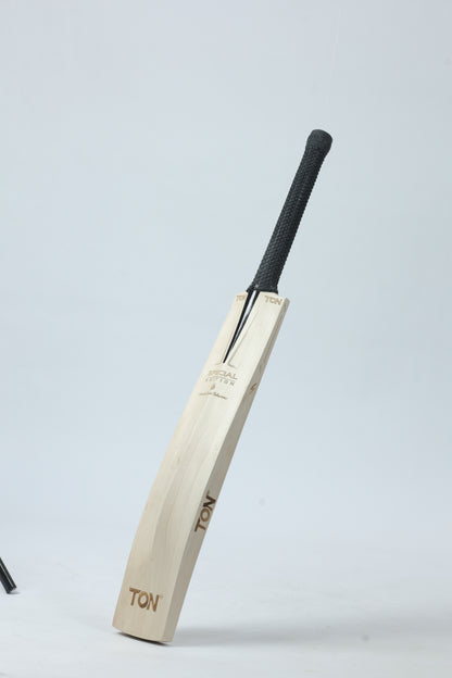 A wooden cricket bat with a black textured grip handle, standing upright against a light gray studio background. The bat features the brand name 'TON' printed on the blade.