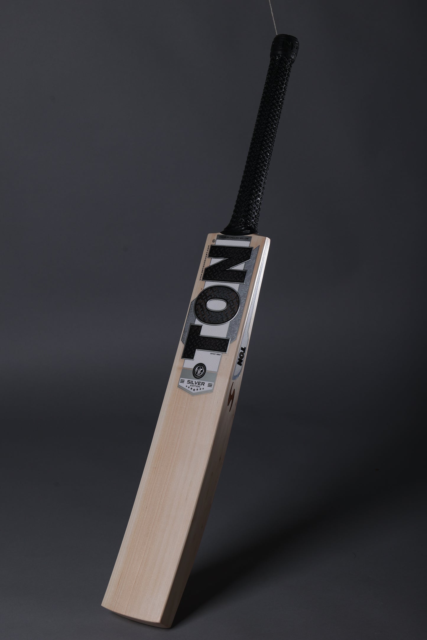 A wooden cricket bat with a black wrapped handle, standing upright against a dark gray studio background. The blade features the brand name 'TON' in bold black letters on a white label.