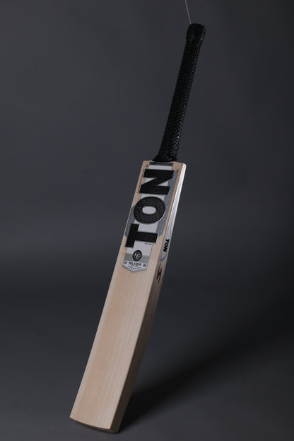 A wooden cricket bat with a black wrapped handle, standing upright against a dark gray studio background. The blade features the brand name 'TON' in bold black letters on a white label.