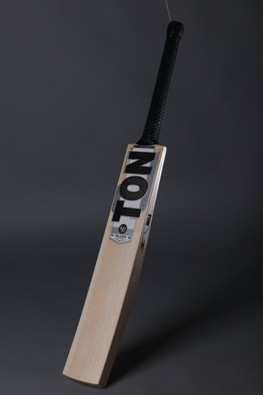 A wooden cricket bat with a black wrapped handle, standing upright against a dark gray studio background. The blade features the brand name 'TON' in bold black letters on a white label.