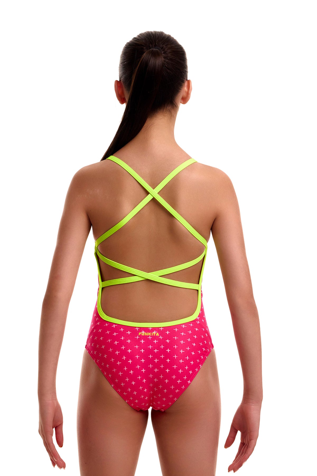 Back view of a young girl wearing the Funkita Strapped In swimsuit in bright pink with a subtle white plus pattern, showing neon lime green criss-cross back straps and a circular cutout.