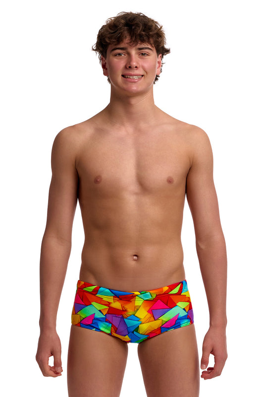 A young male model standing against a solid white background modeling the Funky Trunks "Try The Lot" Boys Sidewinder Trunks, featuring a bright, multi-colored geometric pattern across brief-style swim