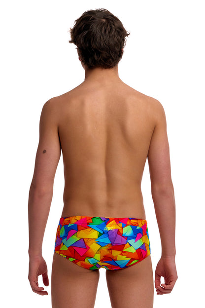 A young male model standing against a solid white background modeling the Funky Trunks "Try The Lot" Boys Sidewinder Trunks, featuring a bright, multi-colored geometric pattern across brief-style swim