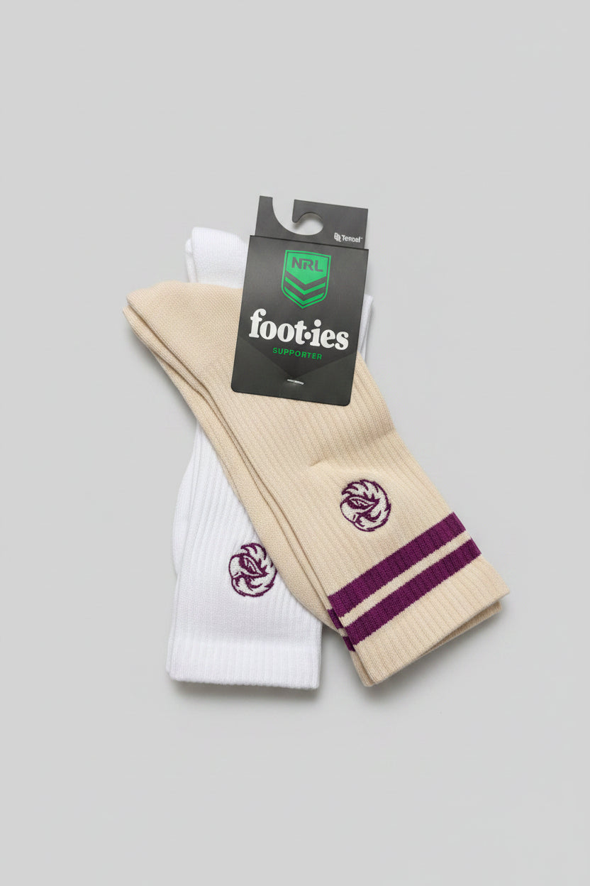 A pair of NRL footies socks, one cream and one white, with purple stripes and logos, resting on a dark patterned carpet. The packaging tag shows the NRL and footies branding.