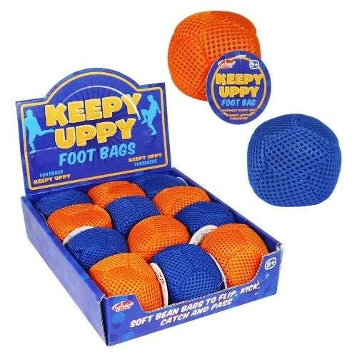 A retail display box of Keepy Uppy Foot Bags, showing several soft mesh bean bags in orange and blue. Two individual bags are shown floating above the box, one orange and one blue, on a plain white background.