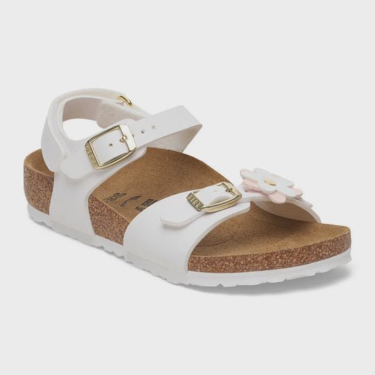 A white leather slide sandal for kids, featuring two adjustable straps with gold-tone buckles, a cork footbed, and a decorative white and pink flower on the side, set against a light gray background.
