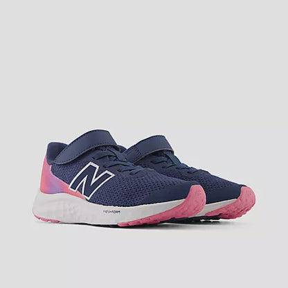 Three-quarter profile view of New Balance Kids Arishi v4 Hook & Loop running shoes in vintage indigo blue, featuring a gradient pink heel, white Fresh Foam midsole, signal pink rubber outsole, and white 'N' logo against a light grey background.