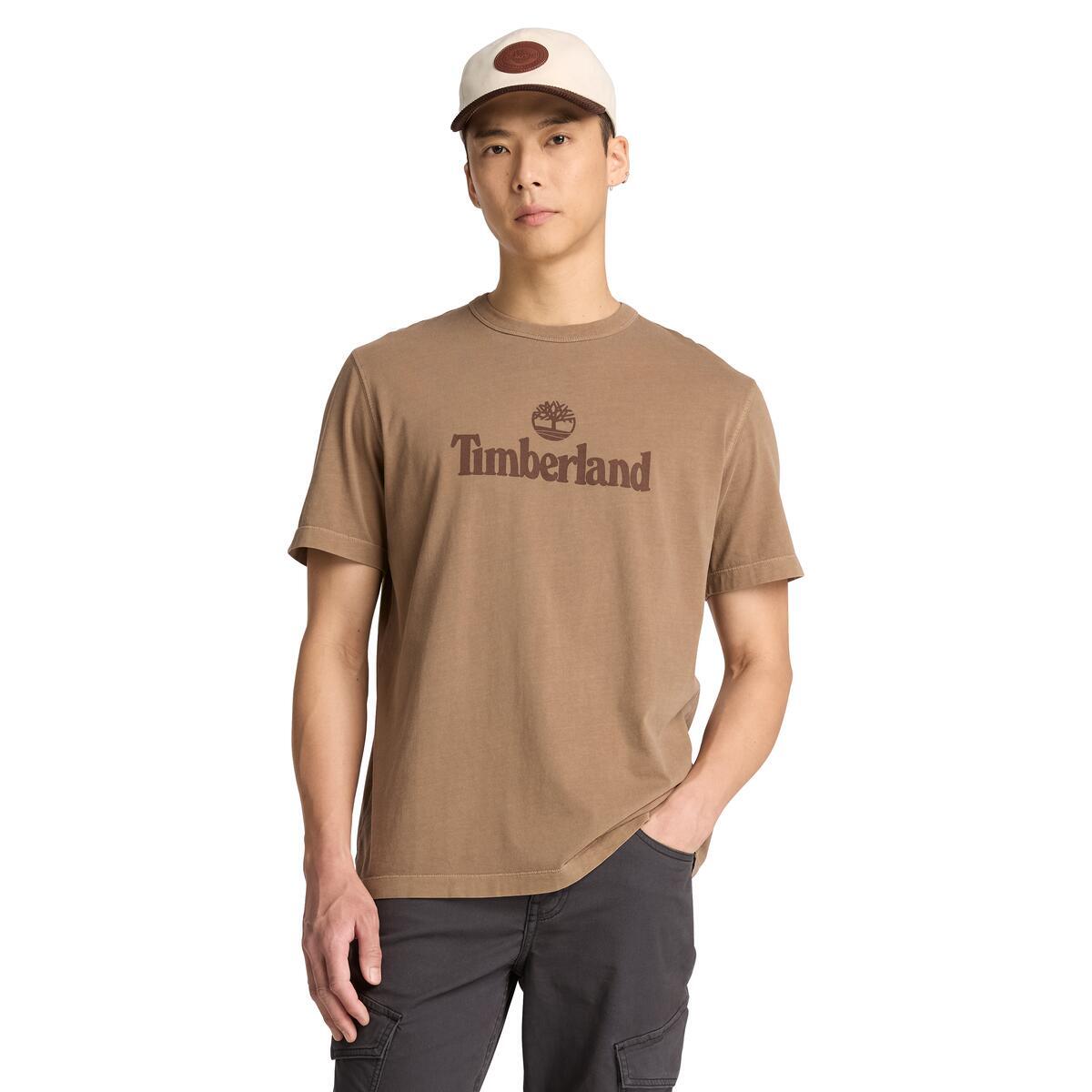 A male wearing a brown short-sleeve crewneck t-shirt with a dark brown Timberland logo printed on the chest.